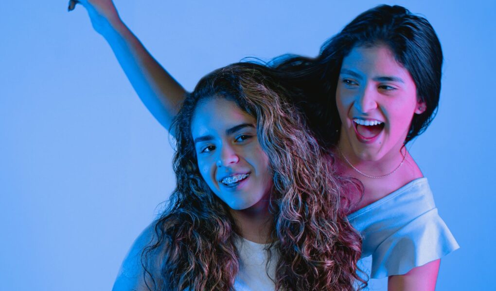 Two young women smiling and posing joyfully in a studio setting with vibrant blue lighting.