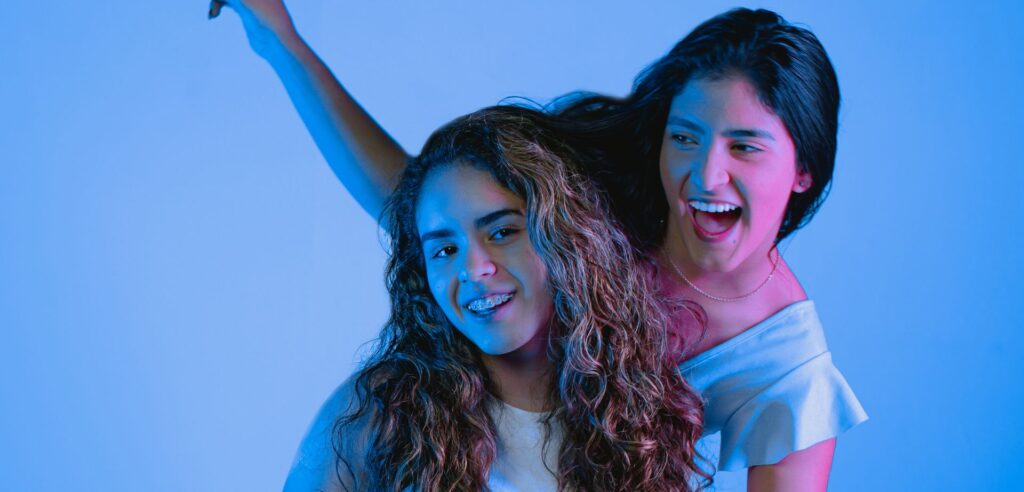 Two young women smiling and posing joyfully in a studio setting with vibrant blue lighting.