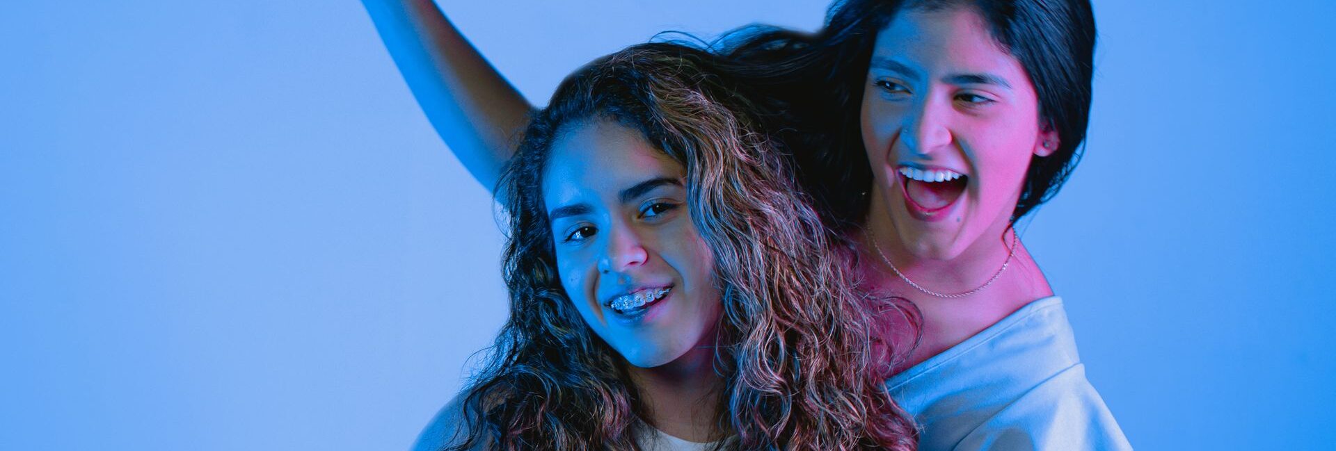 Two young women smiling and posing joyfully in a studio setting with vibrant blue lighting.