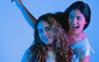Two young women smiling and posing joyfully in a studio setting with vibrant blue lighting.