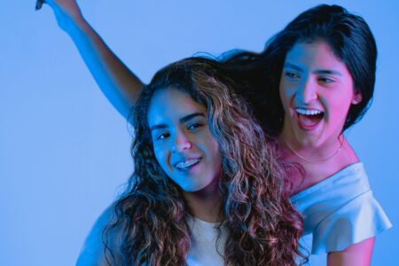 Two young women smiling and posing joyfully in a studio setting with vibrant blue lighting.
